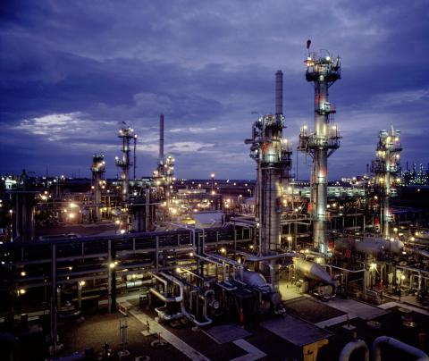 An industrial refinery complex illuminated at dusk. The scene features numerous tall distillation columns, interconnected pipes, and machinery. Bright lights from the facility create a stark contrast against the darkening sky