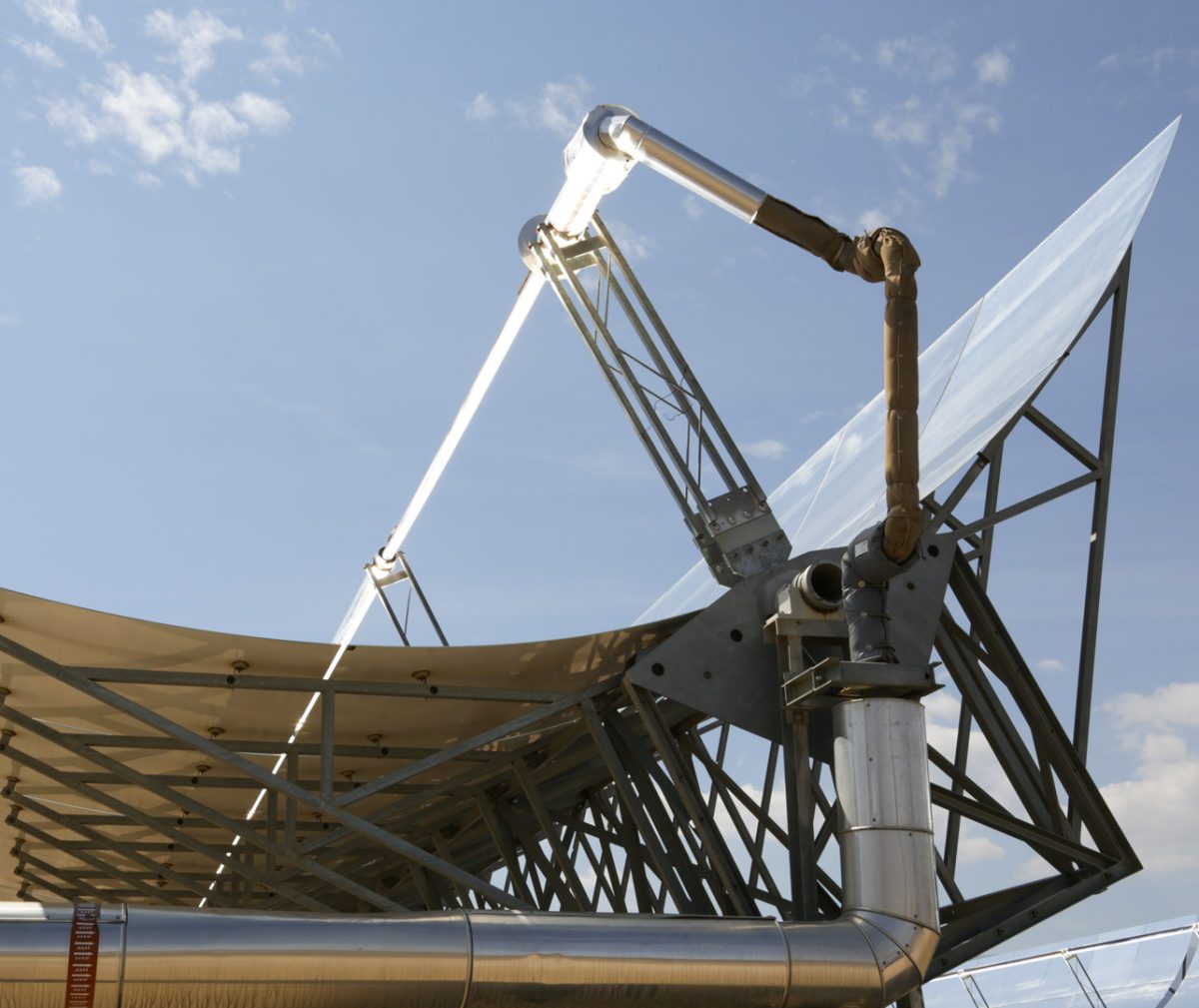 A close-up of a parabolic trough solar collector, a type of solar thermal energy system. The structure features a large, curved reflective surface that concentrates sunlight onto a central receiver tube running along its focal line.