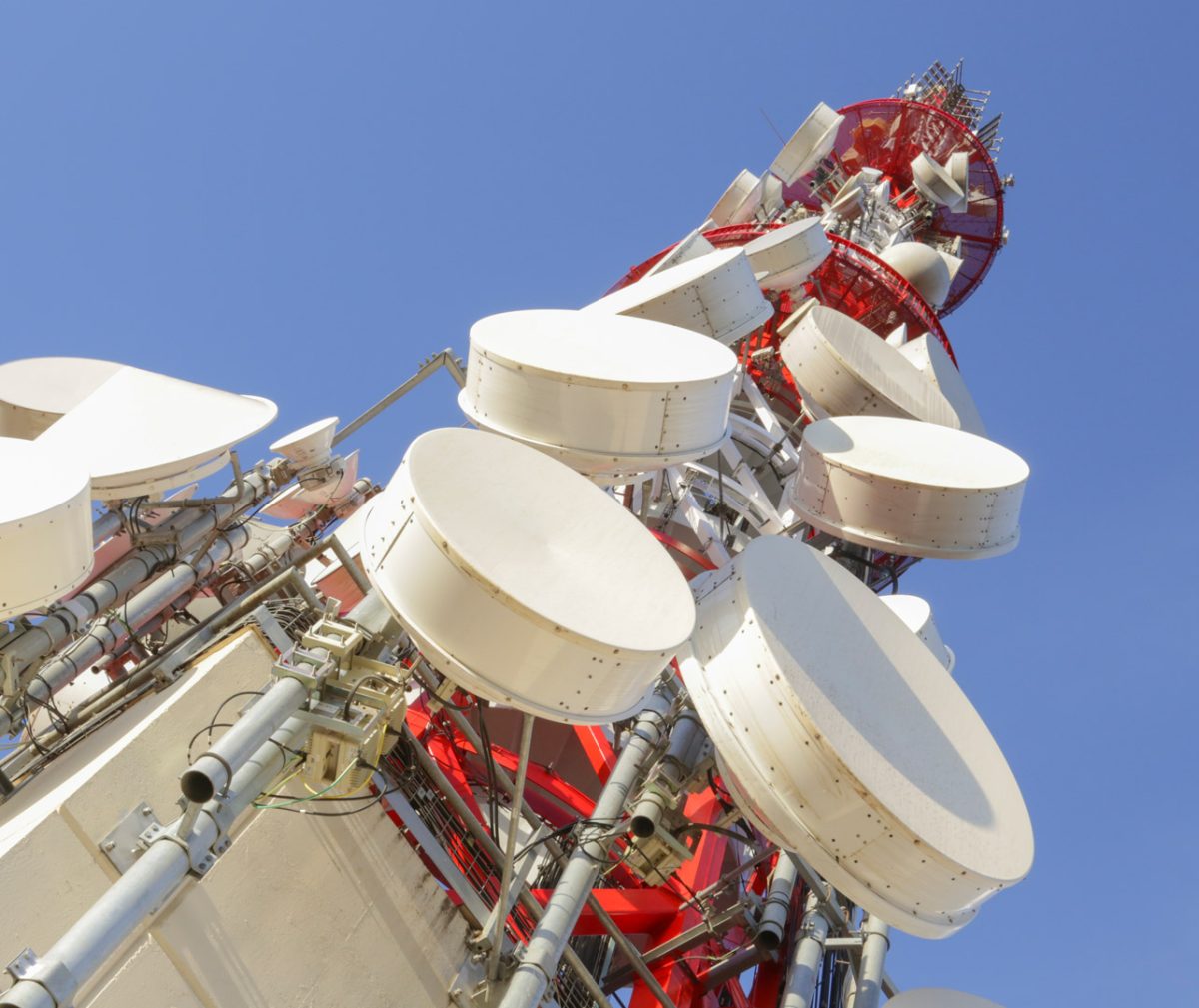 A close-up view of a telecommunications tower, emphasising its various large, round antenna dishes. The tower structure is painted red and white, and the image is taken from a low angle looking up, highlighting the height and complexity of the tower.
