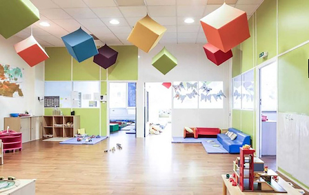 A colourful preschool reception, with blue, red, green, yellow and purple cubes hanging from the ceiling. The furniture is colourful and playful, with toys scattered throughout the room.