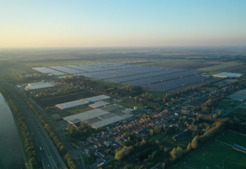 A large solar farm set within a vast, open landscape. The solar panels are arranged in extensive, orderly rows covering a significant area of land. Surrounding the solar farm are patches of green vegetation and open fields.