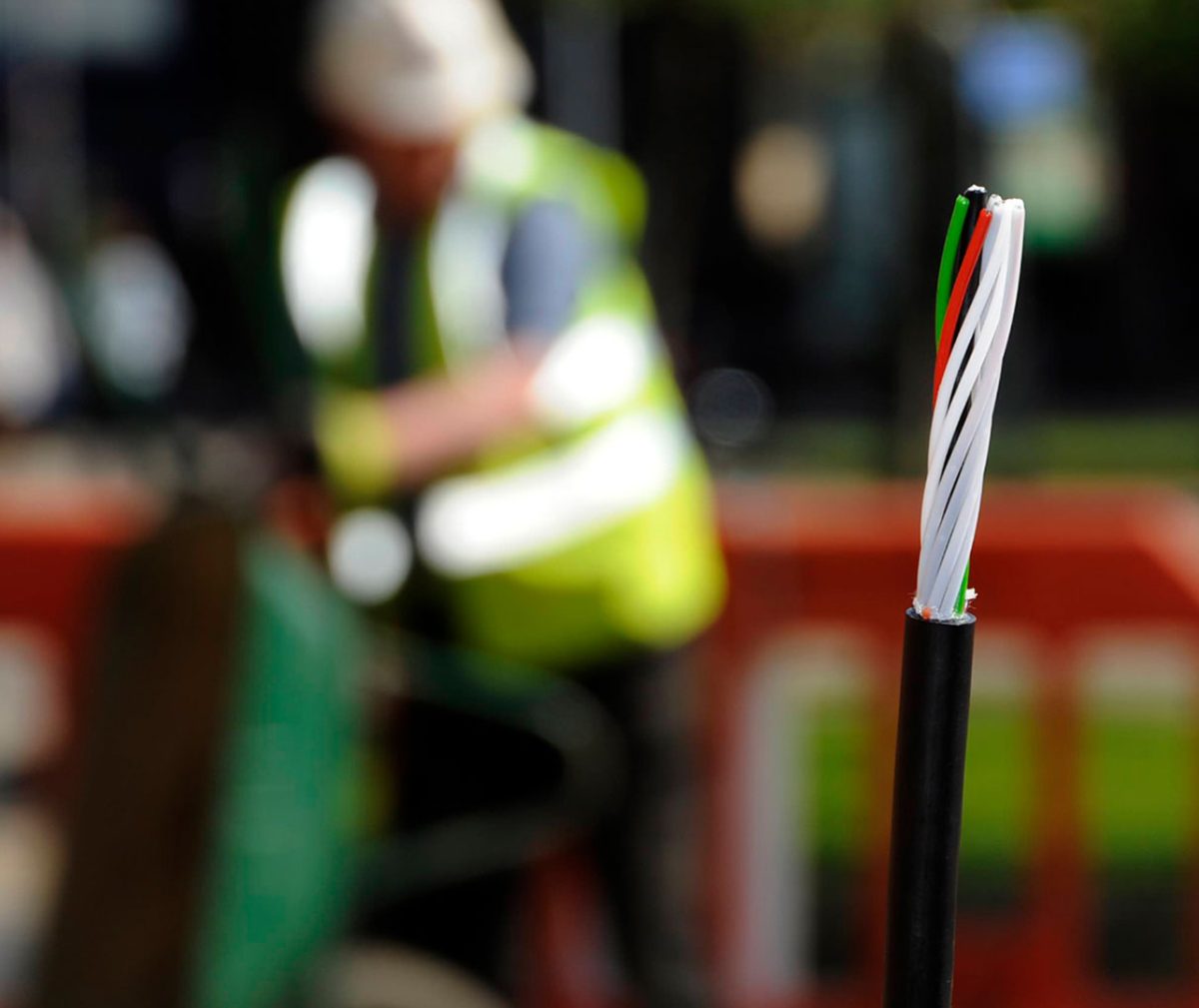 A close-up of a fiber optic cable with several internal wires exposed, each coated in different colours including white, red, green, and black. In the blurred background, a construction worker wearing a high-visibility vest and a hard hat is engaged in work.