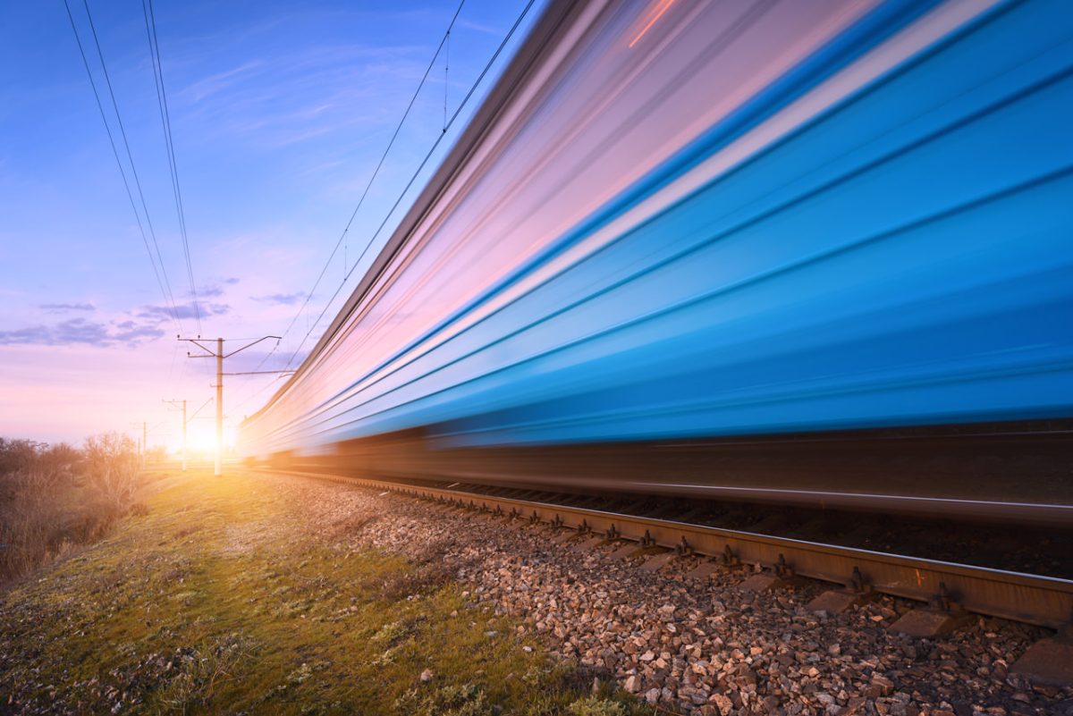 A high-speed train in motion, captured with a blurred effect to convey its rapid movement. The train, painted in shades of blue, is traveling along a set of railway tracks through a rural landscape at sunset.