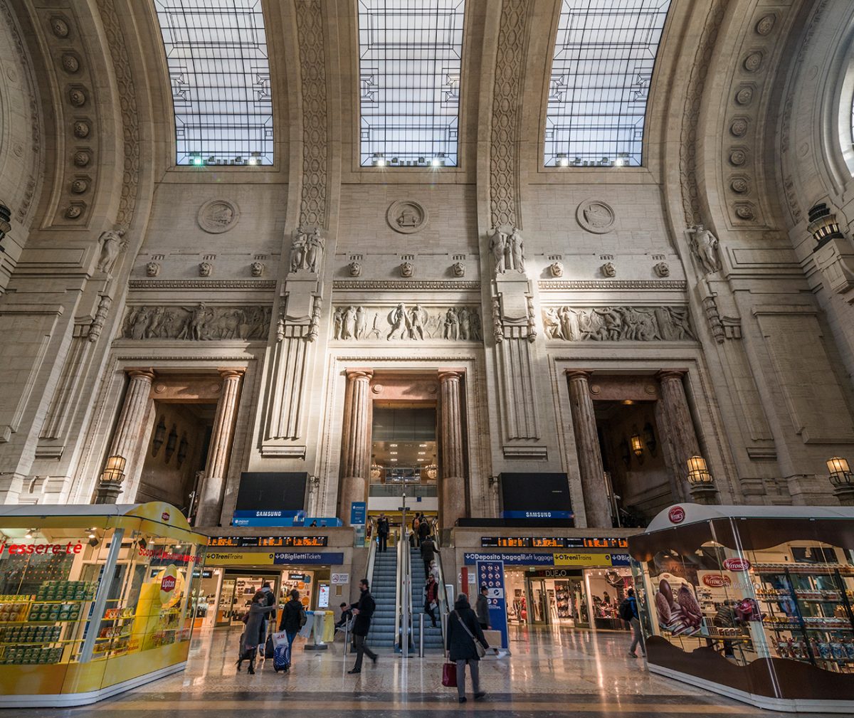The inside of a grand train station in Italy, with an intricate architecture featuring classical statues and a glass ceiling.
