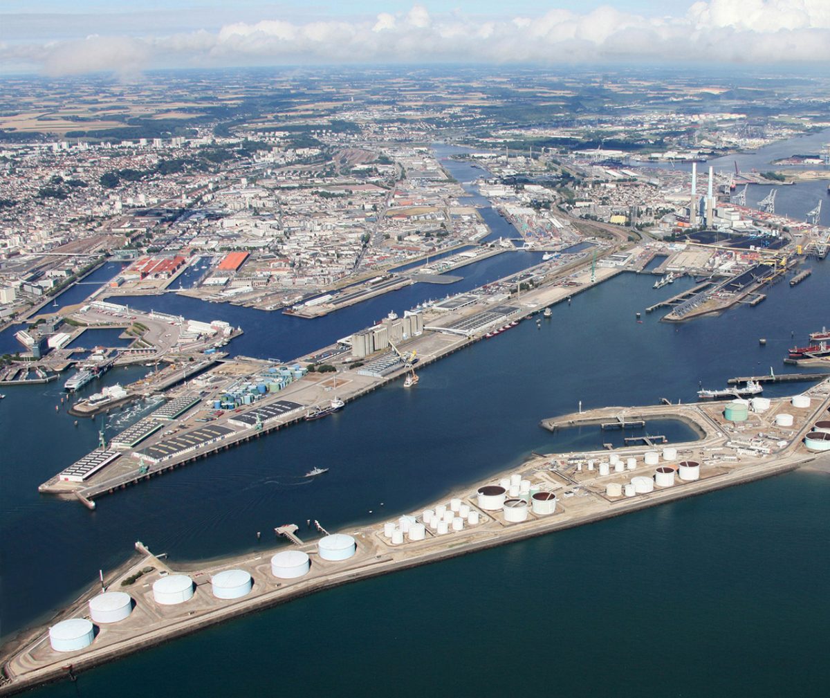 An aerial view of an expansive industrial port complex. The port features numerous docks, warehouses, and storage tanks along its waterfront. Large cylindrical tanks, likely used for storing petroleum or other bulk liquids, are prominently visible in the foreground.