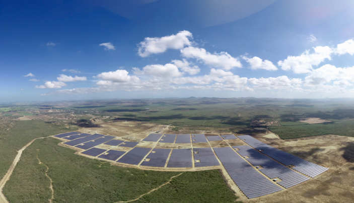 A large solar farm set within a vast, open landscape. The solar panels are arranged in extensive, orderly rows covering a significant area of land. Surrounding the solar farm are patches of green vegetation and open fields.
