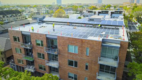 A multi-storey brick residential building with a large array of solar panels on its flat rooftop, with workers in safety gear working on solar panel installation.