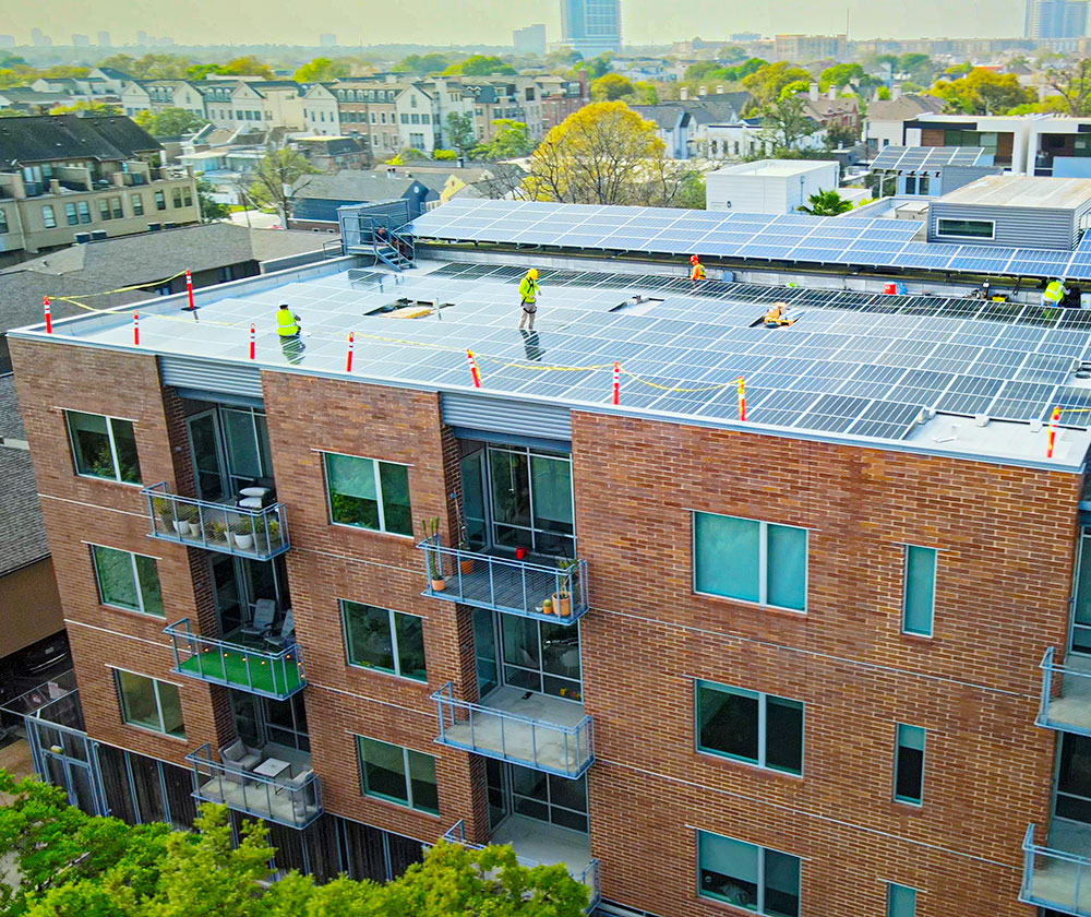 A multi-storey brick residential building with a large array of solar panels on its flat rooftop, with workers in safety gear working on solar panel installation.