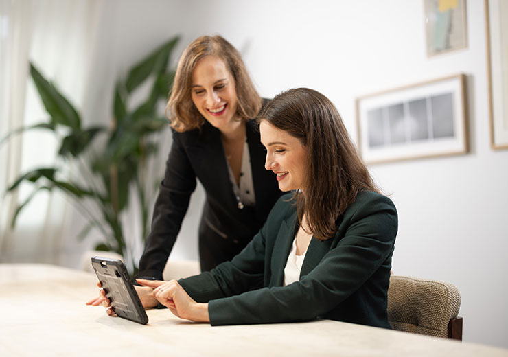 Two women dressed smartly smiling while looking at a tablet in a professional setting.