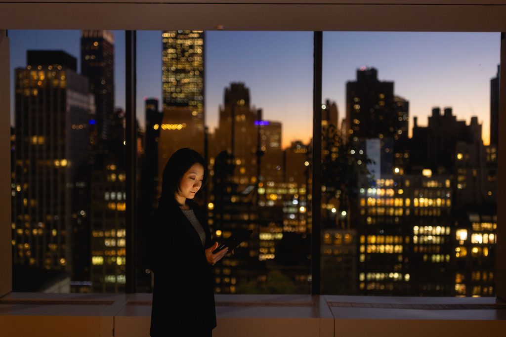 Image of woman in an office building against large windows showing the city skyline, checking her phone. Outside, the sun is setting.